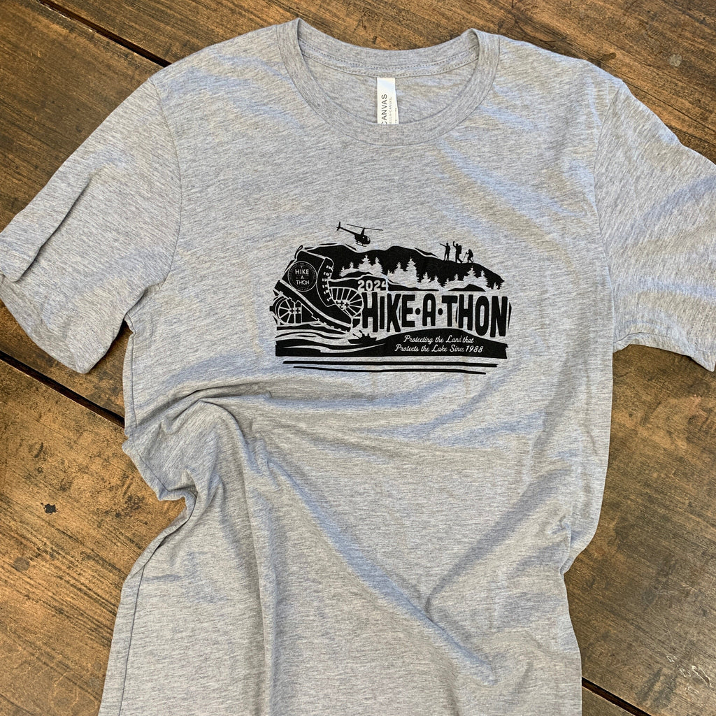 Hike-A-Thon graphic printed in black ink on a heathered gray t-shirt. Logo shows a hiking boot with the Hike-A-Thon logo, next to "2024 Hike-A-Thon" and the tagline "Protecting the Land that Protects the Lake Since 1988". The background is of a mountain silhouetted with trees, hikers, and a helicopter flying overhead, and people in a canoe underneath.