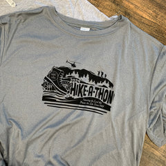 Hike-A-Thon graphic printed in black ink on a darker steel gray t-shirt. Logo shows a hiking boot with the Hike-A-Thon logo, next to "2024 Hike-A-Thon" and the tagline "Protecting the Land that Protects the Lake Since 1988". The background is of a mountain silhouetted with trees, hikers, and a helicopter flying overhead, and people in a canoe underneath.