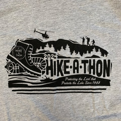 Hike-A-Thon graphic printed in black ink on a heathered gray t-shirt. Logo shows a hiking boot with the Hike-A-Thon logo, next to "2024 Hike-A-Thon" and the tagline "Protecting the Land that Protects the Lake Since 1988". The background is of a mountain silhouetted with trees, hikers, and a helicopter flying overhead, and people in a canoe underneath.