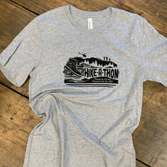 Hike-A-Thon graphic printed in black ink on a heathered gray t-shirt. Logo shows a hiking boot with the Hike-A-Thon logo, next to "2024 Hike-A-Thon" and the tagline "Protecting the Land that Protects the Lake Since 1988". The background is of a mountain silhouetted with trees, hikers, and a helicopter flying overhead, and people in a canoe underneath.