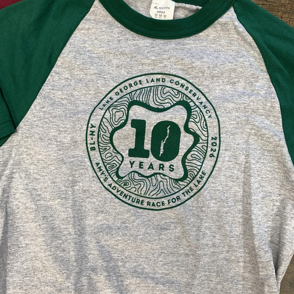 A heather gray and green raglan baseball t-shirt featuring a circular green graphic on the chest that reads '10 YEARS' and 'BL-NY LAKE GEORGE LAND CONSERVANCY 2026 AMY'S ADVENTURE RACE FOR THE LAKE'.