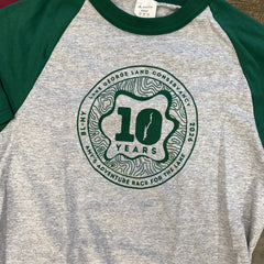 A heather gray and green raglan baseball t-shirt featuring a circular green graphic on the chest that reads '10 YEARS' and 'BL-NY LAKE GEORGE LAND CONSERVANCY 2026 AMY'S ADVENTURE RACE FOR THE LAKE'.