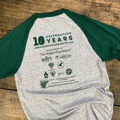 A heather gray and dark green raglan baseball t-shirt laid flat on a rustic wooden surface, showing the back graphic with 'Celebrating 10 Years Amy's Adventure Race for the Lake' and sponsor logos.
