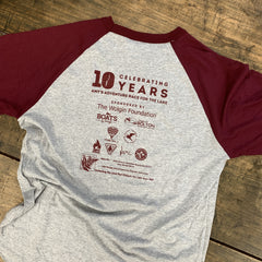A heather gray and maroon raglan baseball t-shirt laid flat on a rustic wooden surface. The front features a maroon graphic print celebrating 10 years of 'Amy's Adventure Race for the Lake' with sponsor logos.