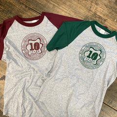 Two raglan-style 10-year baseball t-shirts laid flat on a rustic wooden surface. One shirt has a heather gray body with maroon sleeves and collar, featuring a maroon chest graphic. The other shirt has the same heather gray body with dark green sleeves and collar, featuring a green chest graphic.