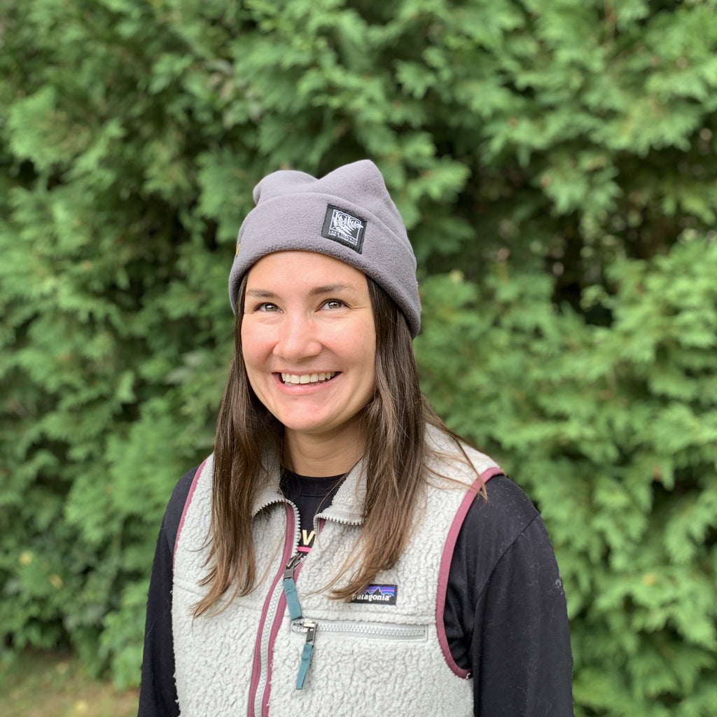 A smiling woman outdoors wearing a gray fleece beanie, a light gray fleece vest over a black shirt, with a blurred green forest background.
