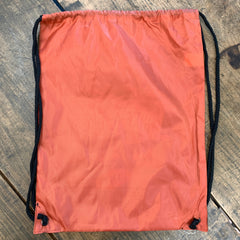Photo shows the back of a rectangular burnt orange tote bag on a wooden table. The bag has two black drawstrings attached to the bottom corners.