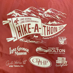 Close-up of the drawstring bag shows a burnt orange background and a cream-colored screen print of a pennant and mountains above and water below. Text says "Lake George Land Conservancy Hike-A-Thon 2026."