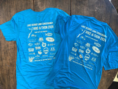 Two aqua blue shirts lay on a brown wooden table. Each has screen printed  words "Lake George Land Conservancy Hike-A-Thon 2026" and "Thank you to our sponsors" in an off-white, and logos of multiple businesses beneath.