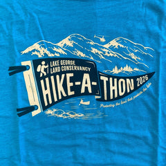 Close-up of an aqua blue shirt with a screen printed image of a pennant in darker blue with the words "Lake George Land Conservancy Hike-A-Thon 2026" in an off-white, with line drawing design of mountains and a helicopter above, and water below with a person in a canoe below. 