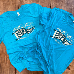 Two aqua blue shirts lay on a brown wooden table. Each has a screen printed image of a pennant in darker blue with the words "Lake George Land Conservancy Hike-A-Thon 2026" in an off-white, with line drawing design of mountains and a helicopter above, and water below with a person in a canoe below. 