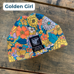 Bold, colorful floral patterned beanie with a black and white LGLC logo embroidered tag on a wooden surface, featuring the style name 'Golden Girl'. 