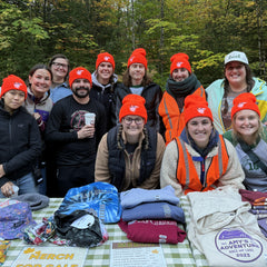 A group of eleven people smiling and posing behind a table outdoors in a forest, with various branded merchandise items displayed on a checkered tablecloth. Most are wearing bright orange beanies featuring an embroidered LGLC logo.