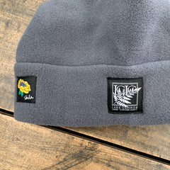 A close-up of a gray fleece beanie resting on a rustic wooden surface. The beanie features a black embroidered patch on the left with a yellow flower and the word 'skida' in white script, and a larger black patch on the right with a white fern design and the text 'LAKE GEORGE LAND CONSERVANCY'.