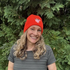 A smiling woman with blonde curly hair wearing a bright orange knit beanie with a white and blue embroidered logo, standing outdoors in front of green evergreen foliage.