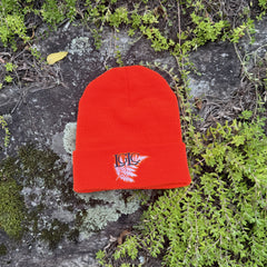 A bright orange knit beanie hat with a folded cuff, featuring an embroidered logo of the letters 'LGLC' in blue over a fern leaf in white. The beanie is photographed flat on a natural mossy rock surface surrounded by green plants.
