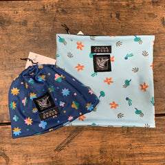 Blue beanie type hat with multi-color stars and rounded shapes lays on a wooden table next to a square neck warmer, light blue printed with orange, brown and green foliage shapes.