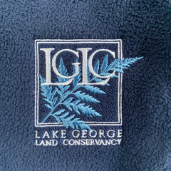 Close-up of embroidered LGLC logo. White stitching used for a double-lined box surrounding the letters "LGLC" and "Lake George Land Conservancy" written beneath. A simple fern is embroidered inside the box, behind the "LGLC" in a bright blue thread.